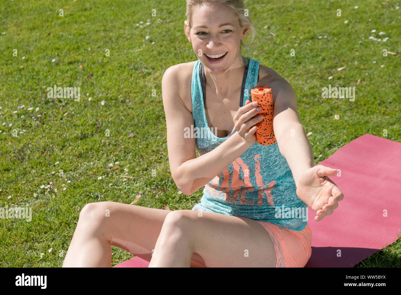 Frau in Trainingskleidung auf rosa matt im Park, Massage Roller am Arm Stockfoto