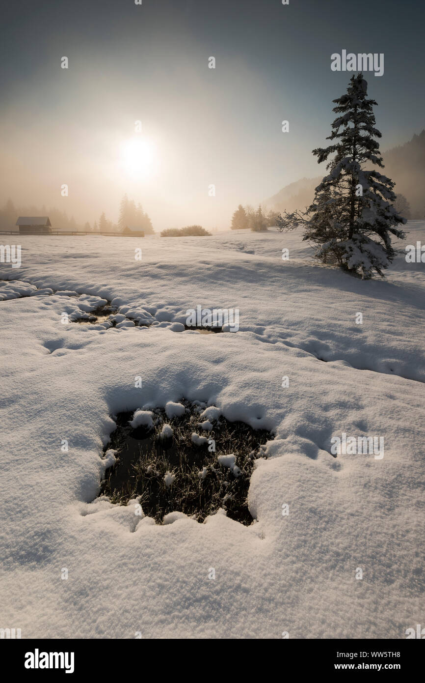 Ein kleiner Wasserlauf im Schnee - Winter Landschaft nahe Garmisch Partenkirchen abgedeckt. Im Hintergrund die aufgehende Sonne am Morgen Nebel mit Holzhütten. Stockfoto