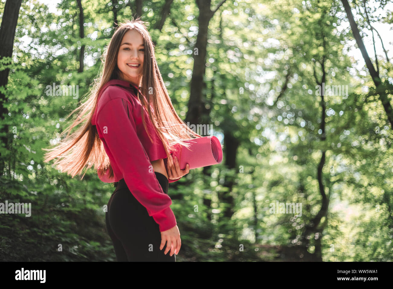 7/8 Bild einer attraktiven Athlet Frau mit flatternden Haare in ein sportliches Sweatshirt mit Kapuze mit einem Sport Matte auf einem Waldweg. Stockfoto