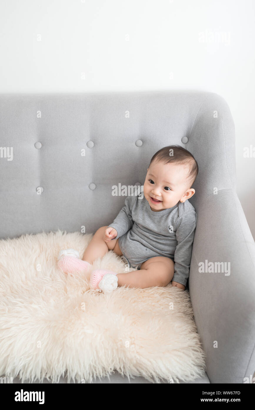 Closeup Portrait von cute adorable Lächeln Lachen, Baby Mädchen mit schwarzen Augen sitzen auf einem Sofa weg von der Kamera suchen, natürliche Fenster Licht, lifestyl Stockfoto