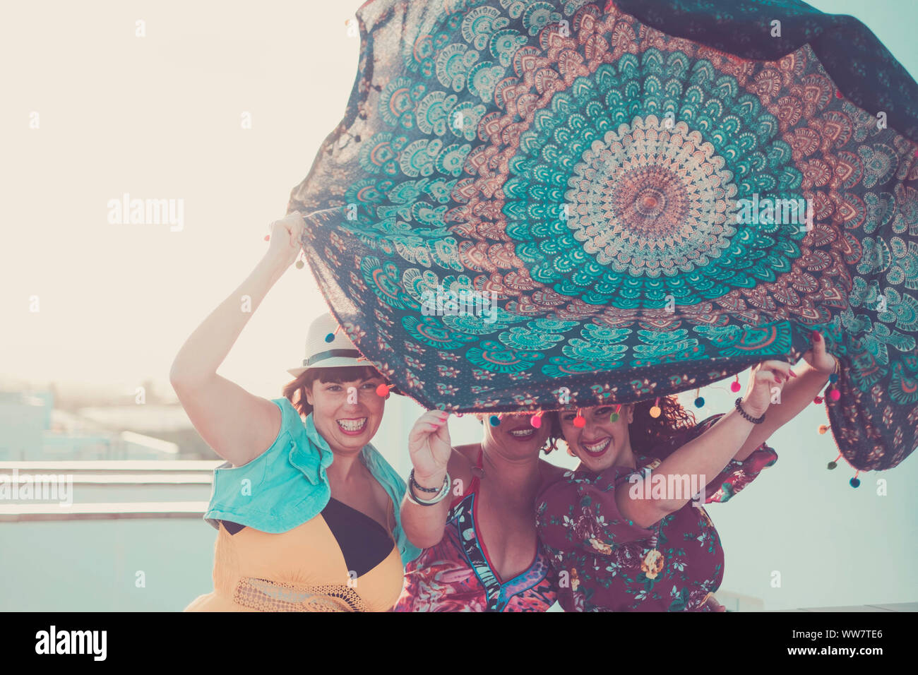 Drei junge schöne weibliche Damen nehmen ein Mandala Textil über die Köpfe. Spielen mit dem Wind in Aktivität im Freien. Sommer Konzept mit Hintergrundbeleuchtung Stockfoto