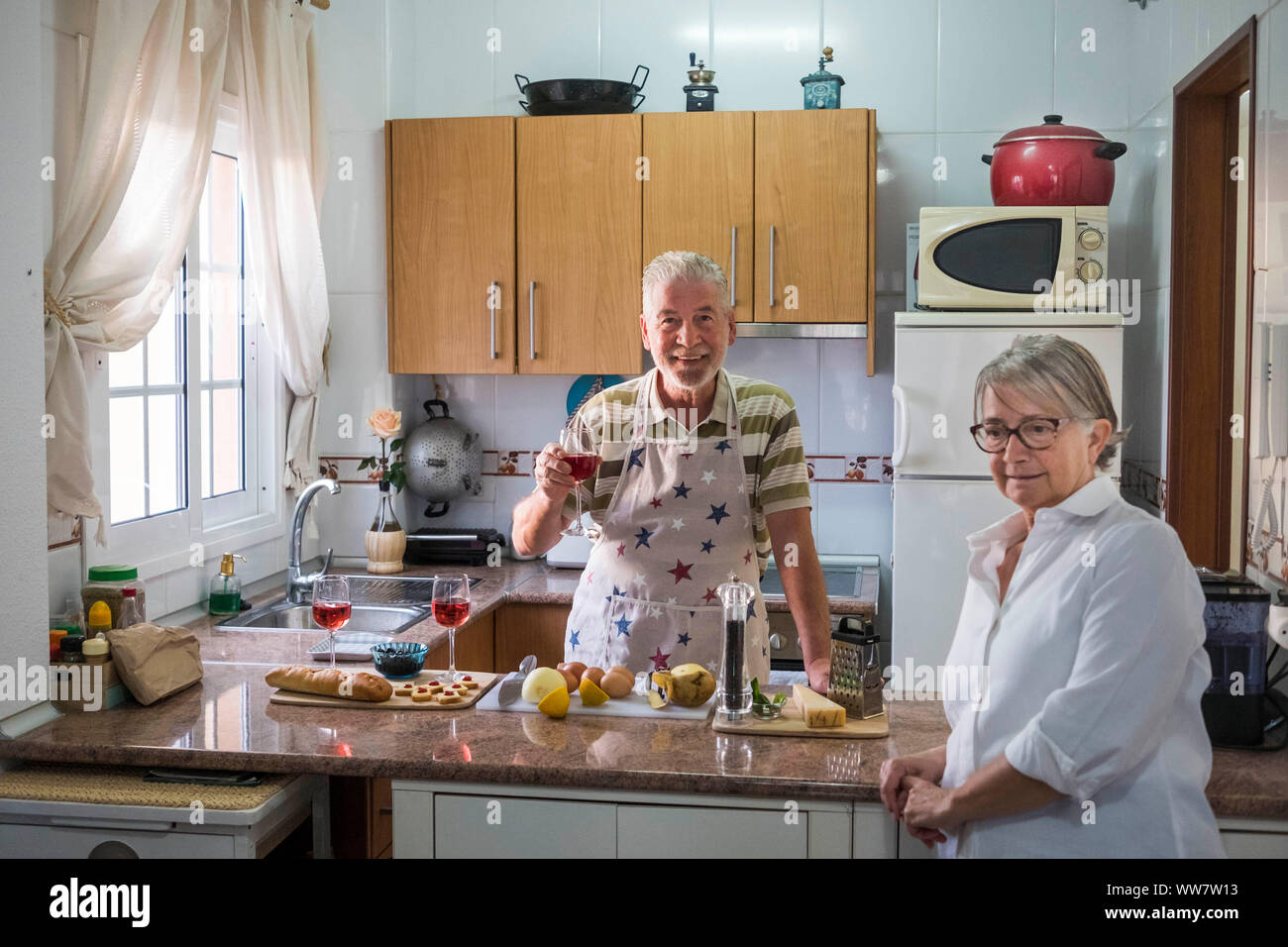 Ein erwachsener älterer Mann Wein zu trinken und dabei etwas für einen Cocktail mit ihrer Frau zu Essen vorbereiten. Altenheim arbeiten in einem üblichen Küche. Licht vom Fenster. Stockfoto