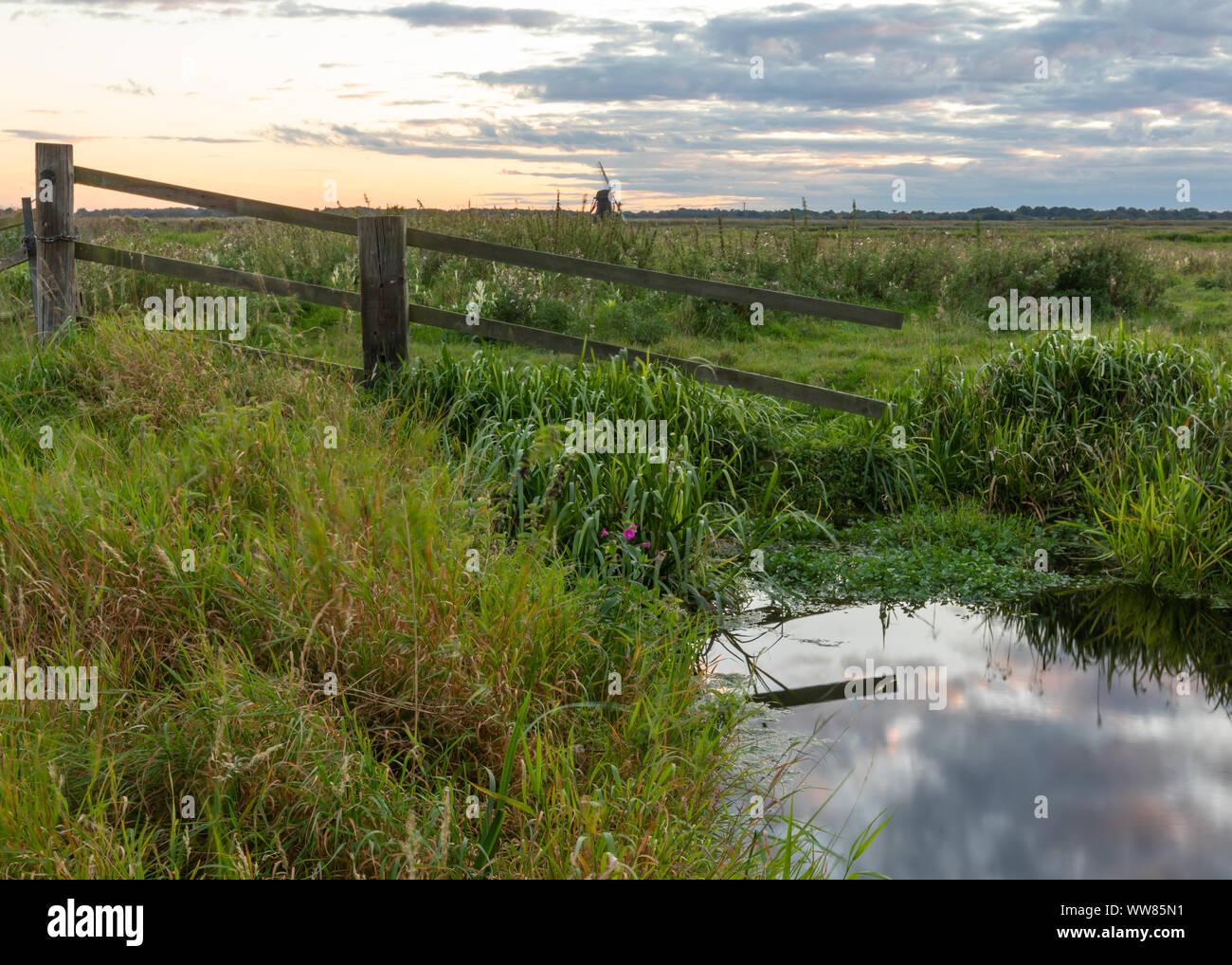 Herringfleet Mühle bei Dämmerung in der Ferne durch ein defektes Zaun auf dem sumpfigen Breite gesehen mit dem Himmel in einem Pool von Wasser in den Vordergrund wider Stockfoto