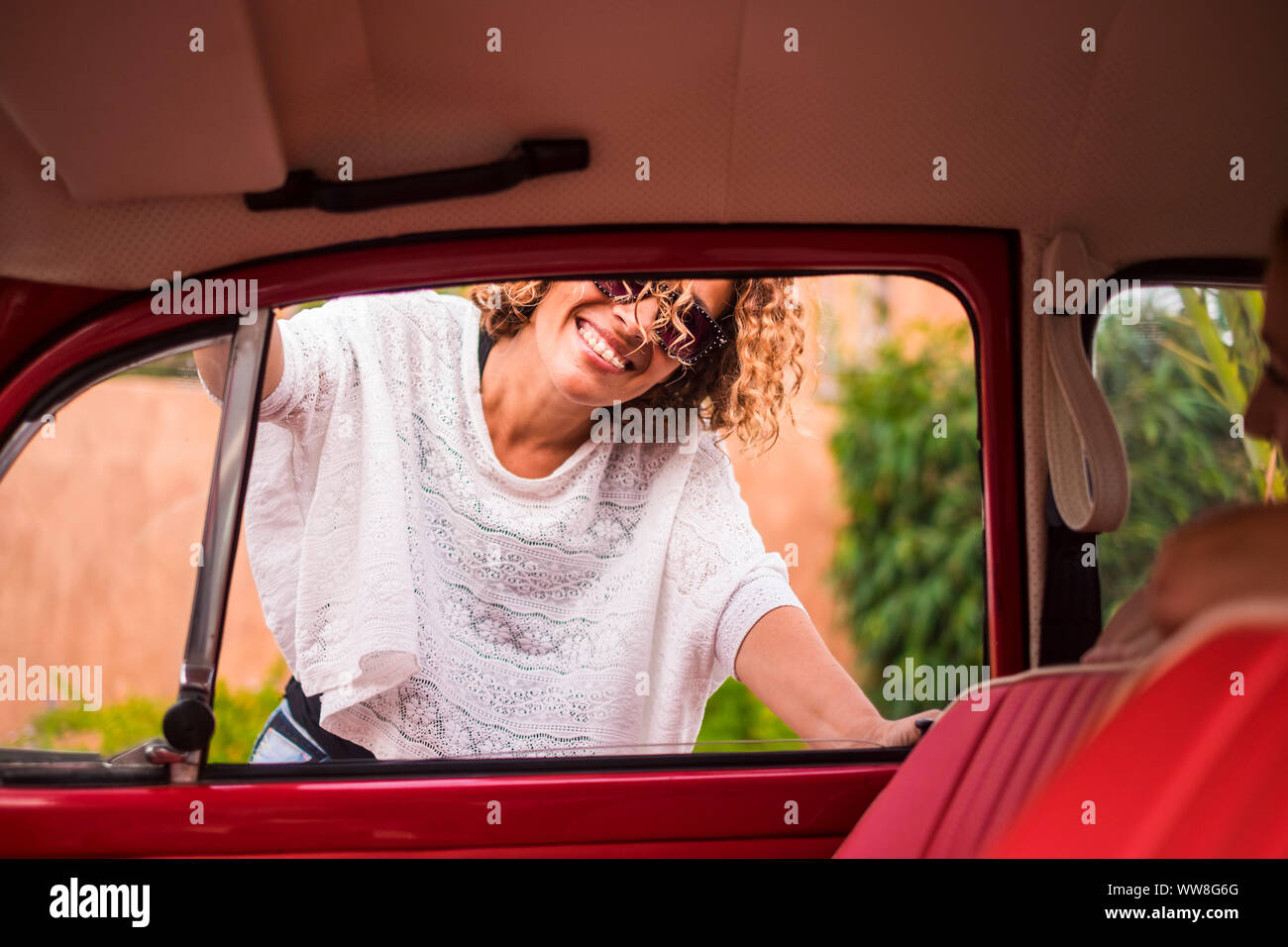 Fröhliche und schöne kaukasischen mittleren Alter curly blonde Frau Lächeln und den Outdoor Freizeitaktivitäten Aktivität in der Nähe von ihrem roten vintage Classic Auto genießen, bereit zu reisen, um die Tag leben und Stockfoto