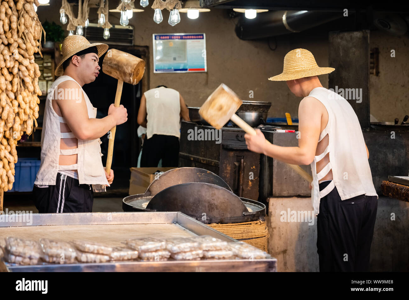 Xian, China - Juli 2019: Zwei Männer schlagen hart mit großen holzhämmern Getreide, das bei der Herstellung von süßen Snack für den Verkauf auf dem str eingesetzt werden zu knacken Stockfoto