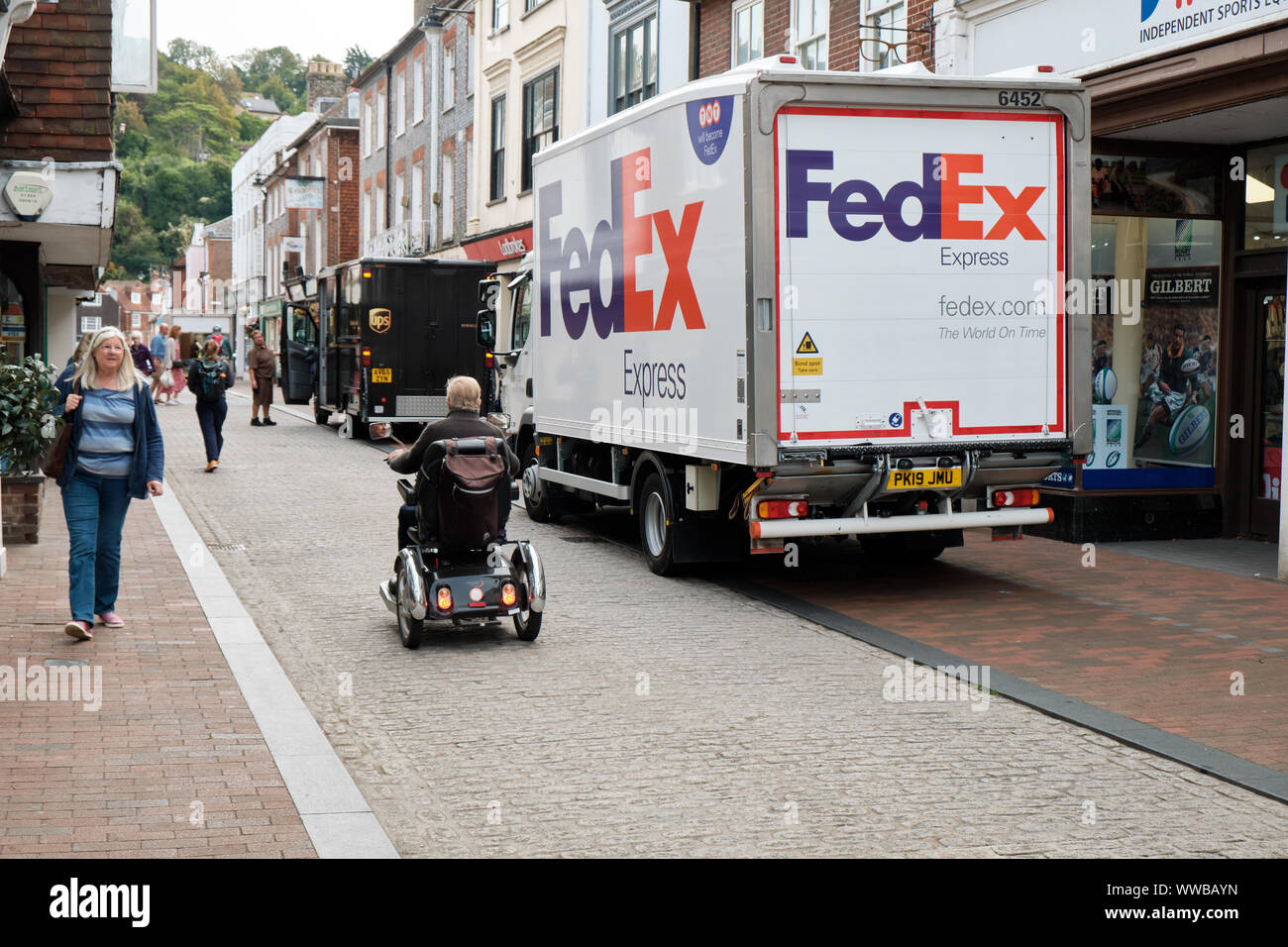Fedex und UPS LKW illegal in der Fußgängerzone der Stadt geparkt, während eine Person mit eingeschränkter Mobilität auf Straße mit Kopfsteinpflaster geht. Lewes GROSSBRITANNIEN Stockfoto