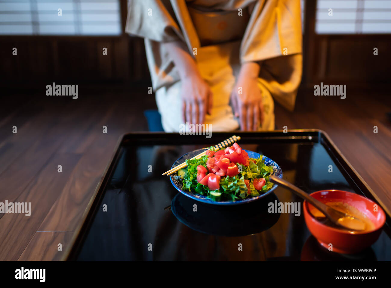 Junge Frau im Kimono seiza an traditionellen japanischen Haus oder Ryokan restaurant Zimmer durch die Tabelle mit Salat Teller und Stäbchen sitzen, Shoji Schiebetüren p Stockfoto