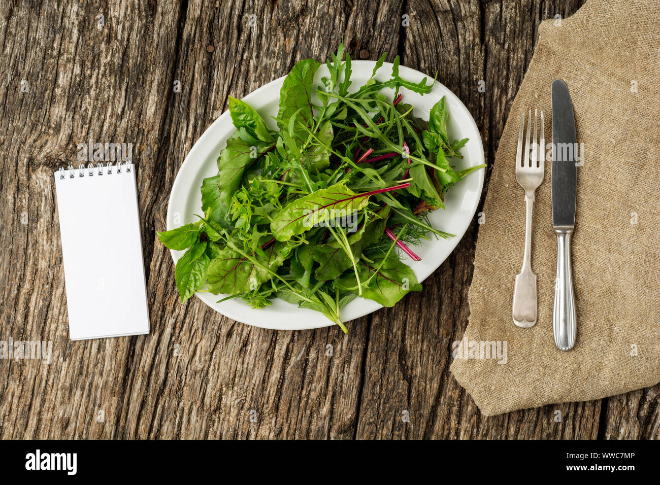 Frische Salatteller mit gemischtem grünen (Rucola, mesclun, papiermache) auf dunklem Hintergrund mit Besteck und Notepad. Gesundes Essen. Grüne Mahlzeit. Flach Stockfoto