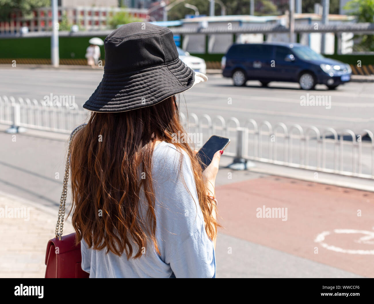 Eine chinesische Frau in einem Sonnenhut steht auf Bürgersteig und hält ein Smartphone. Portrait Foto von hinten, das Gesicht ist nicht sichtbar und der Hintergrund ist unscharf Stockfoto