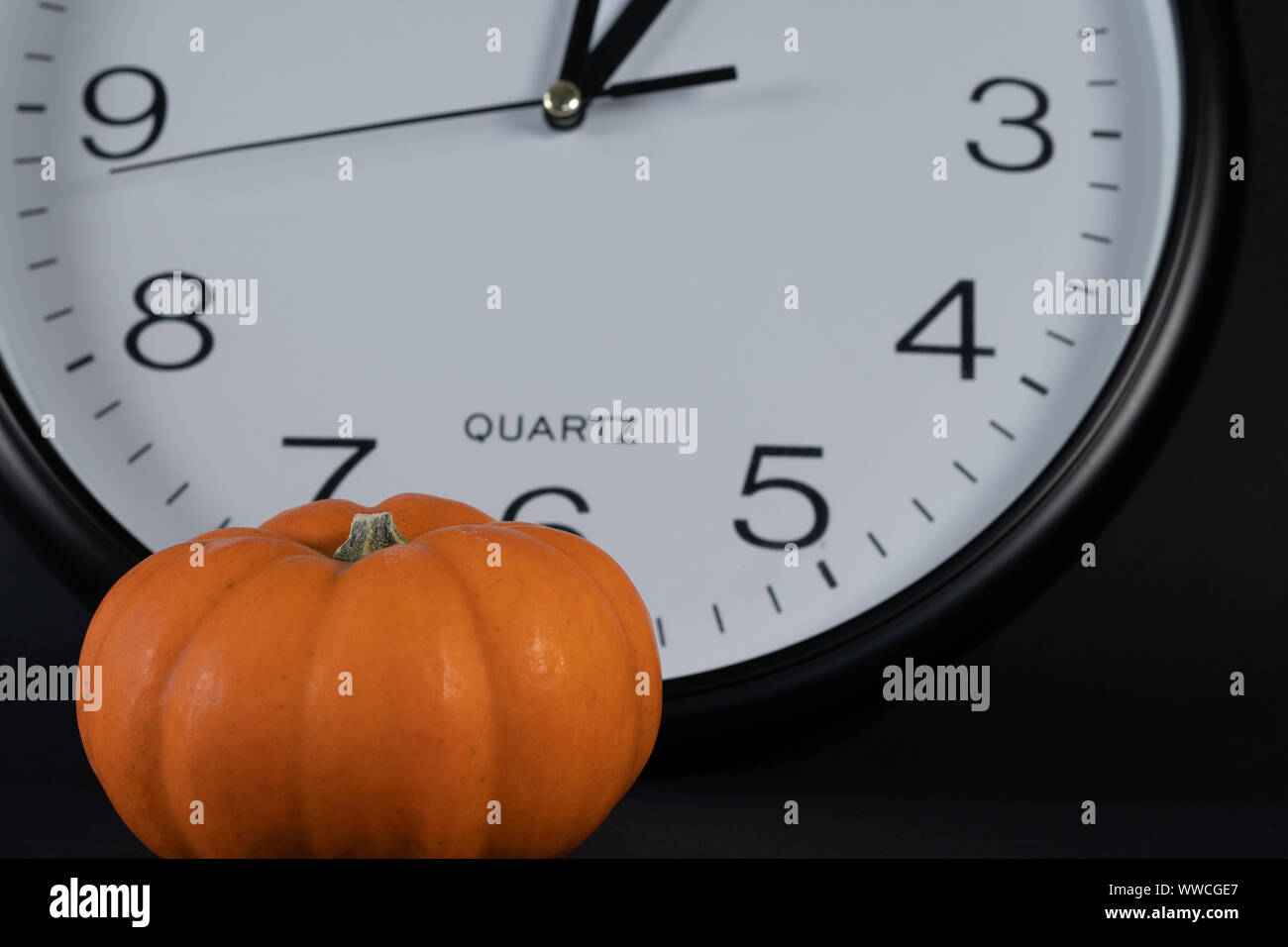 Mini Kürbis im Vordergrund mit großer weißer Uhr auf schwarzem Hintergrund bei 1 Uhr. Konzept für Kürbis Zeit oder Sommerzeit, Nahaufnahme. Stockfoto