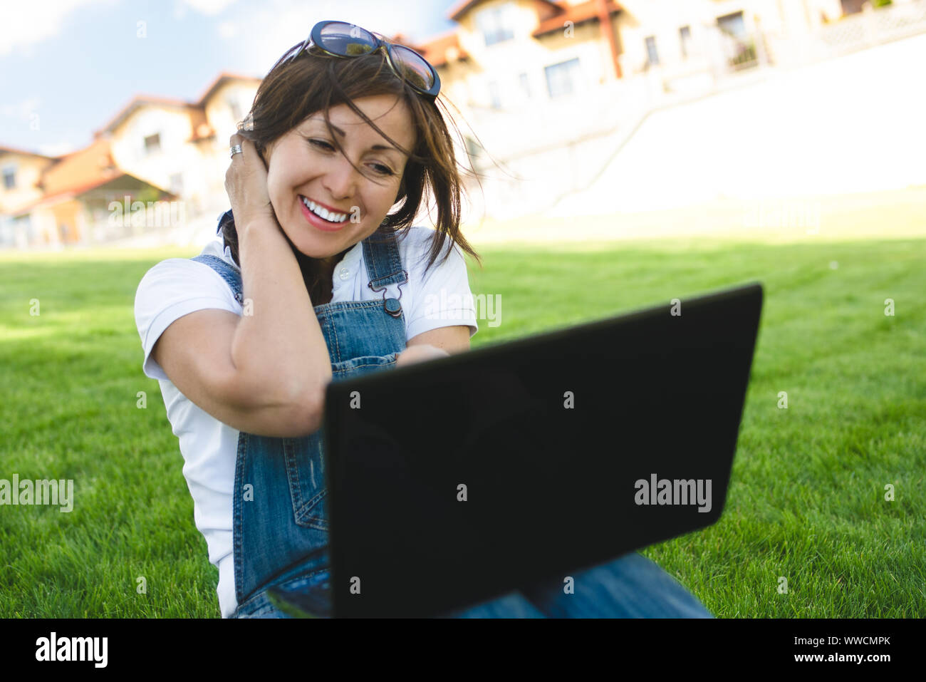 Weibliche erhält freudige Emotionen durch Gespräche mit ihren Freunden auf ein Konferenzgespräch aus Notebook, gekleidet in Jeans Overalls sitzen auf dem Rasen im Park Stockfoto