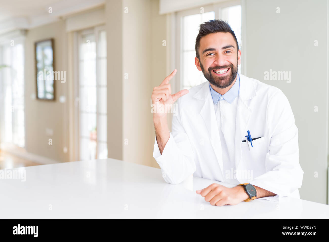 Schöner spanischer Arzt oder Therapeut Mann, medizinische Fell an der Klinik lächelnd und selbstbewusst und deutete mit der Hand tun Größe Schild mit Fingern wh Stockfoto