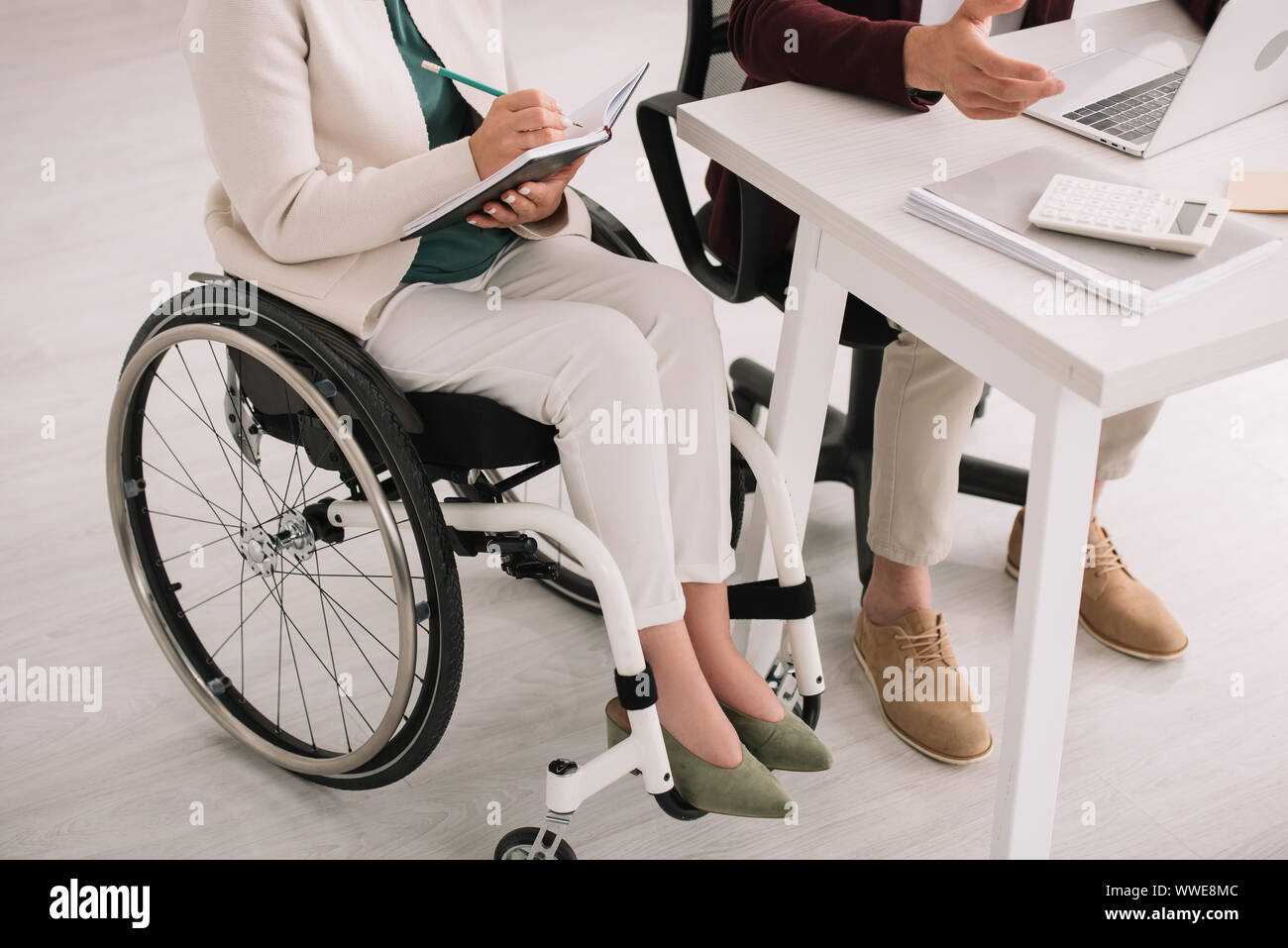 7/8-Ansicht von behinderten Geschäftsfrau schreiben im Notebook beim Sitzen im Rollstuhl in der Nähe von Kollegen Stockfoto