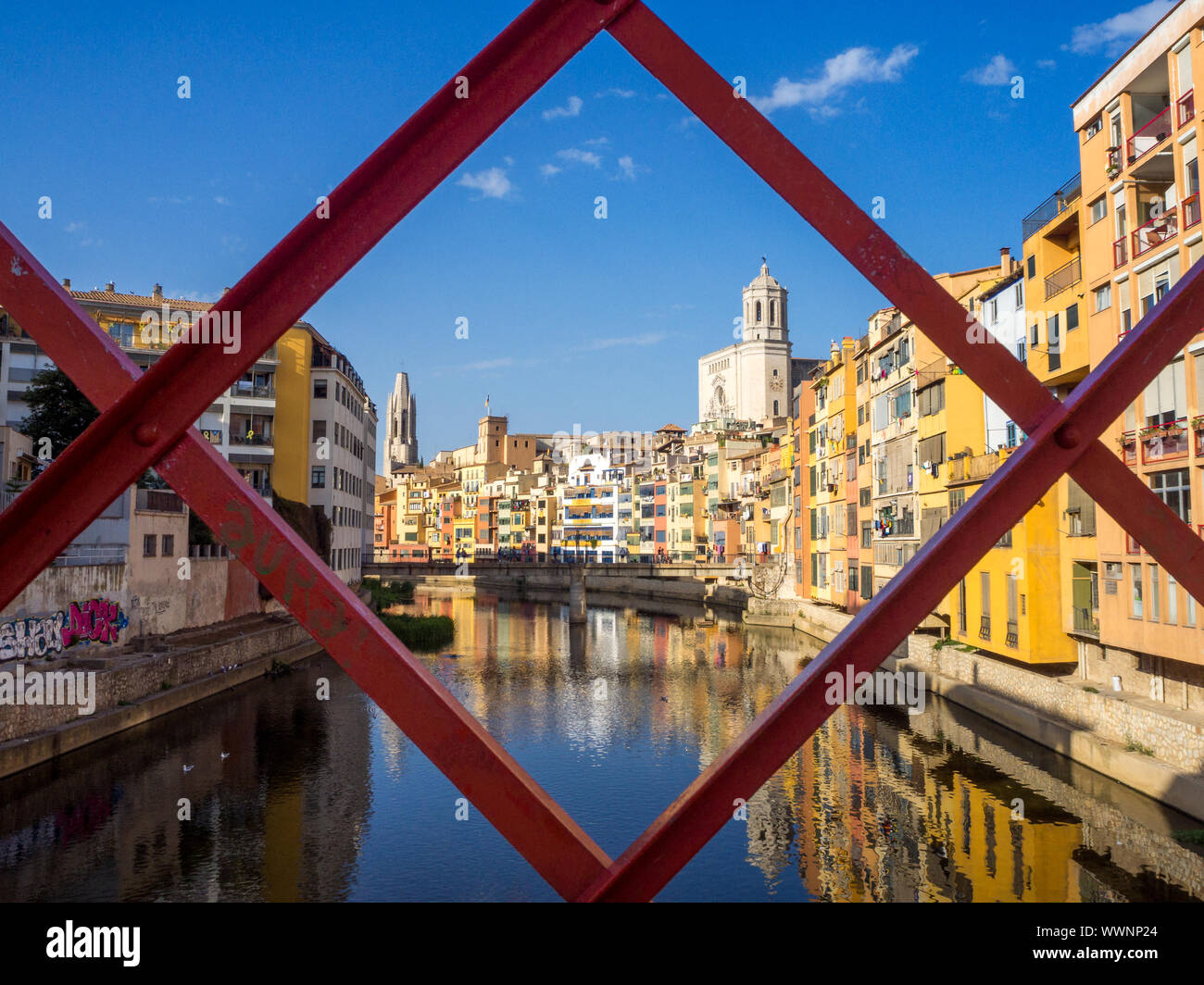 Von Girona typische Skyline Stadtbild durch den Rahmen der roten Eisenbrücke über den Fluss Onyar Fluss mit bunten Häusern auf einem blauen sonnigen Himmel, Kirche o Stockfoto