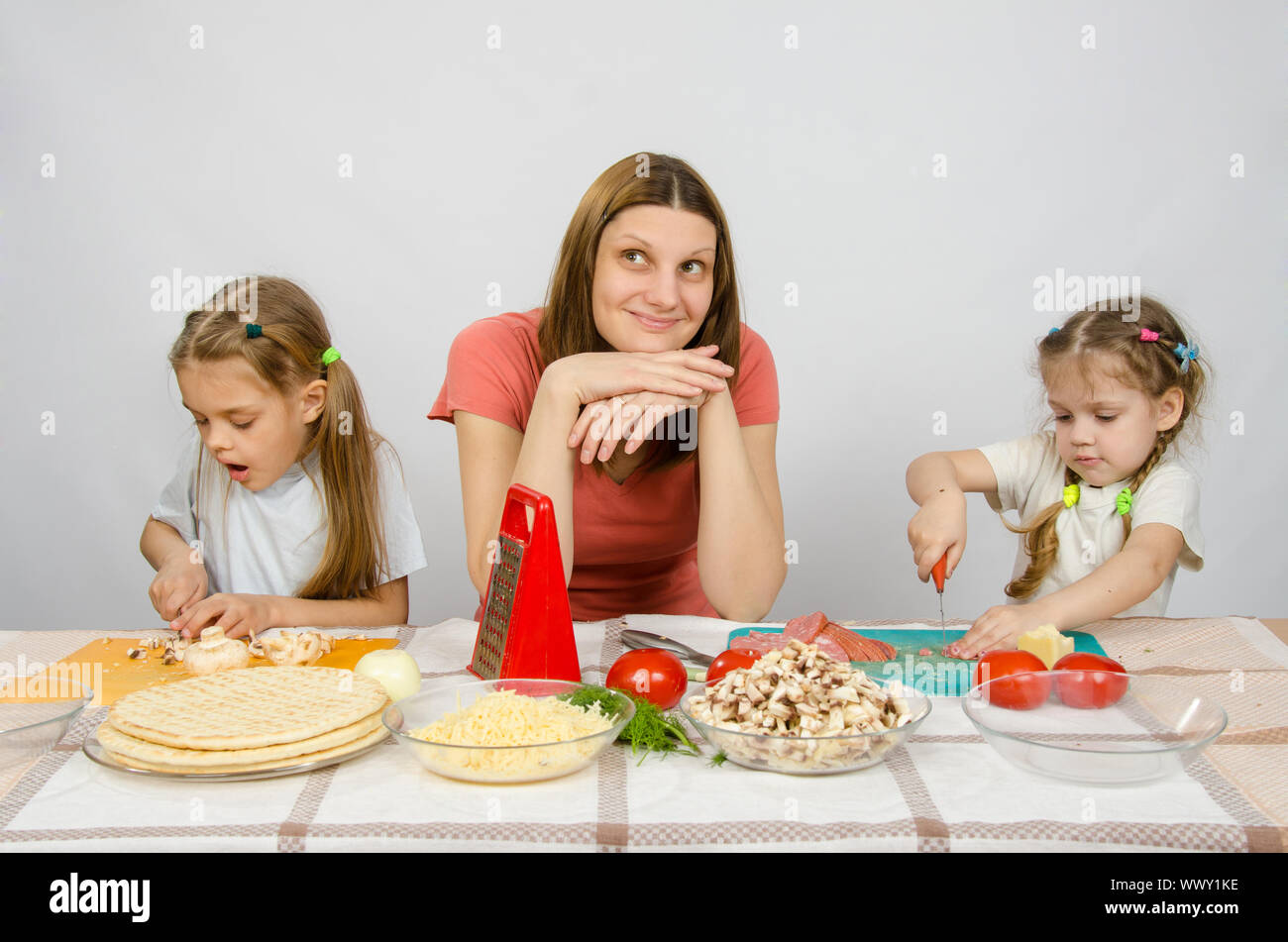 Mutter sitzt am Küchentisch mit einem traumhaften Blick auf zwei kleine Mädchen enge Konzentration geschnittene Produkte Stockfoto