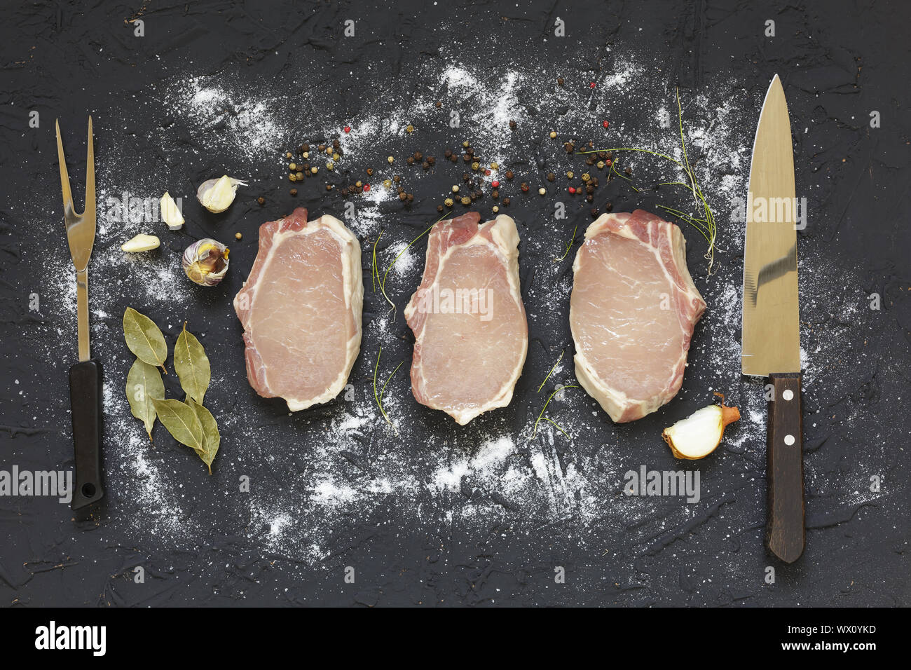 Drei Scheiben, rohes Fleisch, rohes, T-Bone Steak, frische Kräuter, Salz, Knoblauch, Zwiebel, Pfeffer auf einem dunklen Stein Stockfoto