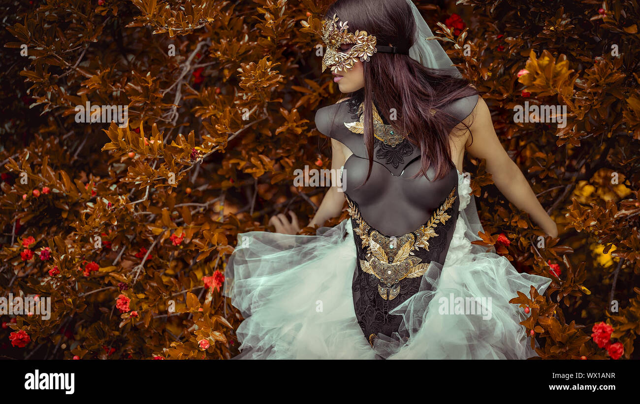 Geheimnisvolle Frau in Weiß gekleidet mit Maske von Blättern in Kupfer und Gold Farbe in einem Garten im Herbst Stockfoto