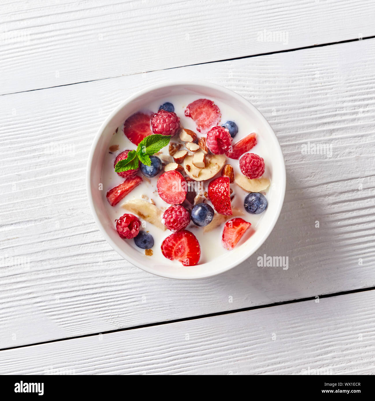 Bunt und lecker bereit Frühstück aus Mais Müsli, Früchte, Milch, Mandeln, Scheiben Banane auf eine Schüssel auf einem weißen Tisch wit Stockfoto