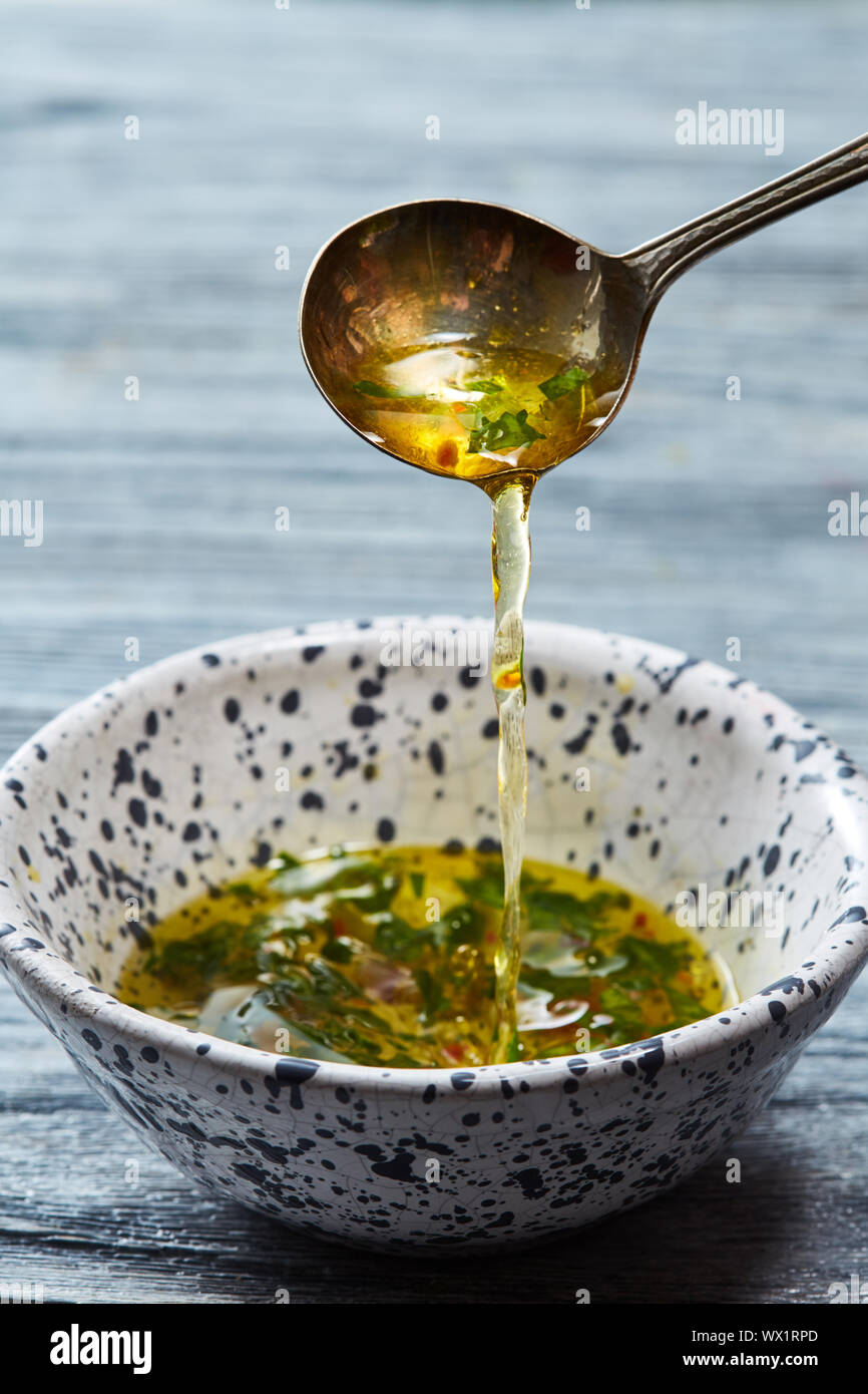 Hausgemachten Salat Dressing mit Olivenöl, Essig, grünen und Gewürze gießen vom Löffel in eine Schüssel auf einem grauen Holztisch. Stockfoto