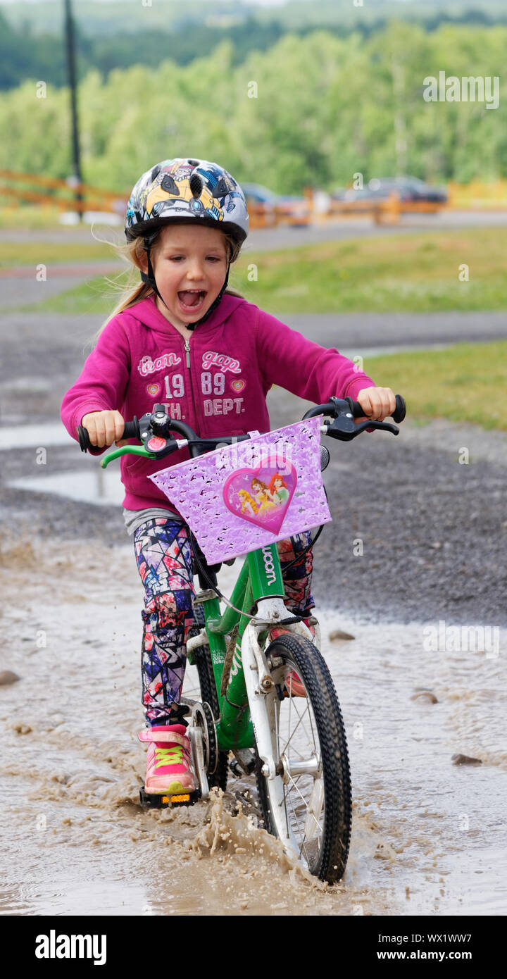 Ein lächelndes Mädchen (5 Jahre alt) reiten ihr Fahrrad durch eine Pfütze Stockfoto