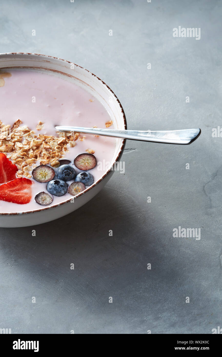Weiße Schüssel mit hausgemachtem Müsli, in Scheiben geschnittene Erdbeeren, Joghurt, Blaubeere, Löffel - natürliche Frühstück auf einem grauen Tabelle. Stockfoto
