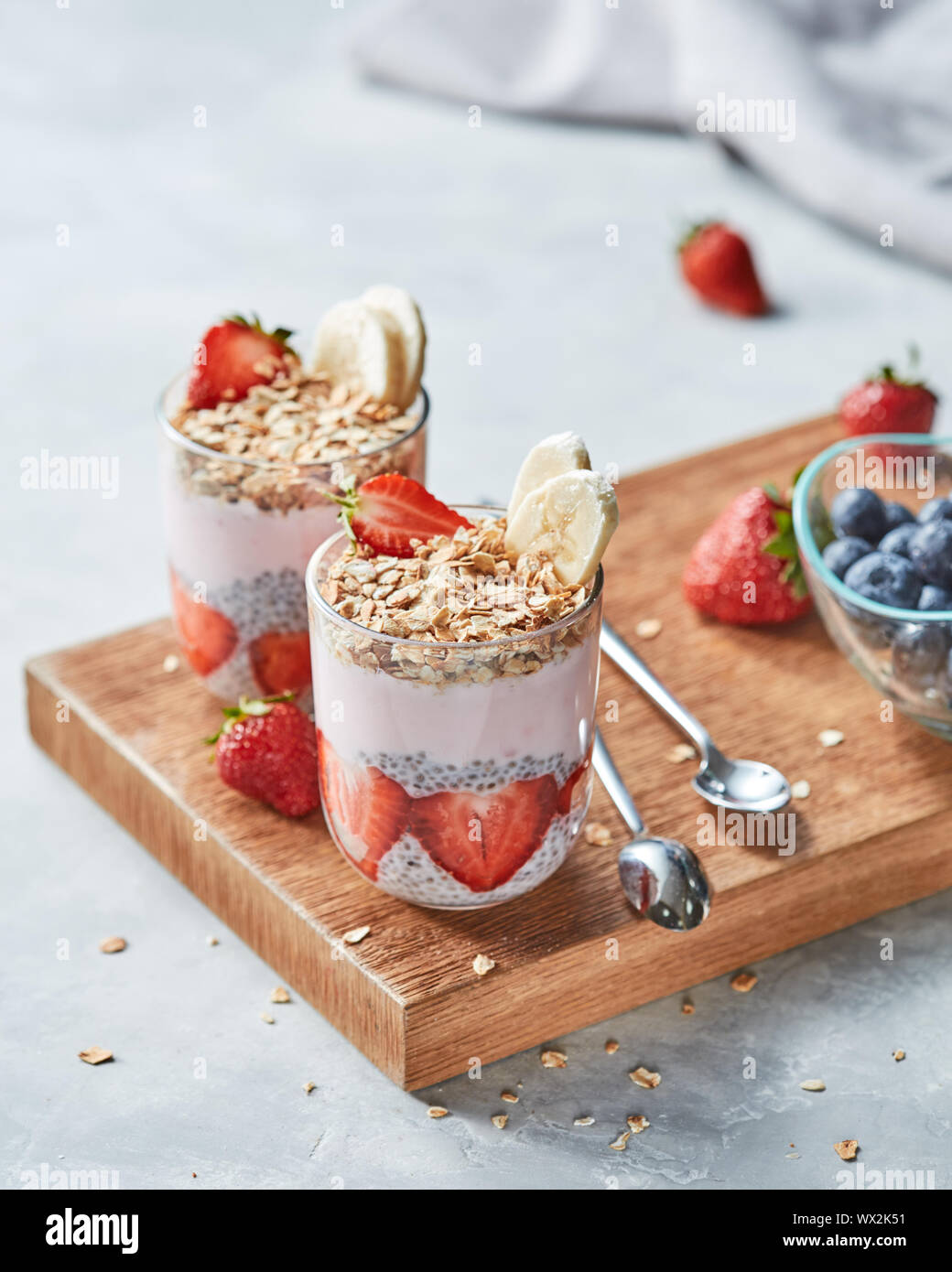 Müsli, Hälften von Erdbeeren, Banane, Chia Samen mit Joghurt in Gläsern auf einem Holzbrett auf grauem Beton, Platz für Tex Stockfoto