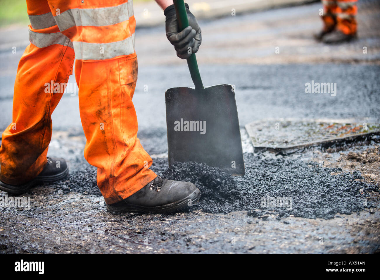 Mann in orange Arbeitshose mit einer Schaufel Verbreitung tar und Splitt auf der Straße erneuert werden Stockfoto