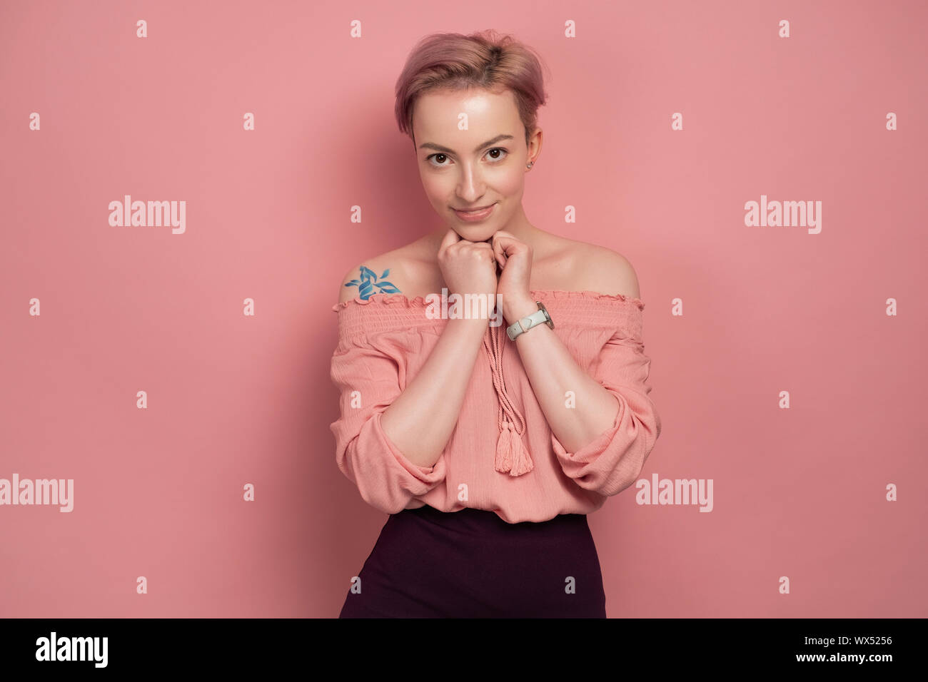 Ein Mädchen mit kurzen rosa Haaren steht auf einem rosa Hintergrund, lächeln süß, mit Blick auf die Kamera, umklammerte ihre Hände an ihre Brust. Stockfoto