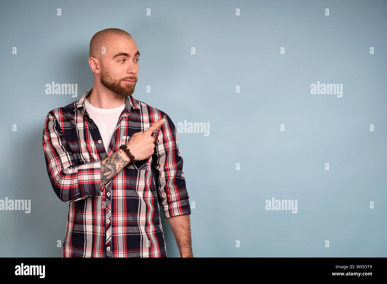 Ein bärtiger Kerl in einem Plaid Shirt steht auf einem blauen Hintergrund und sieht recht und zeigt seinen Zeigefinger an der Seite Stockfoto