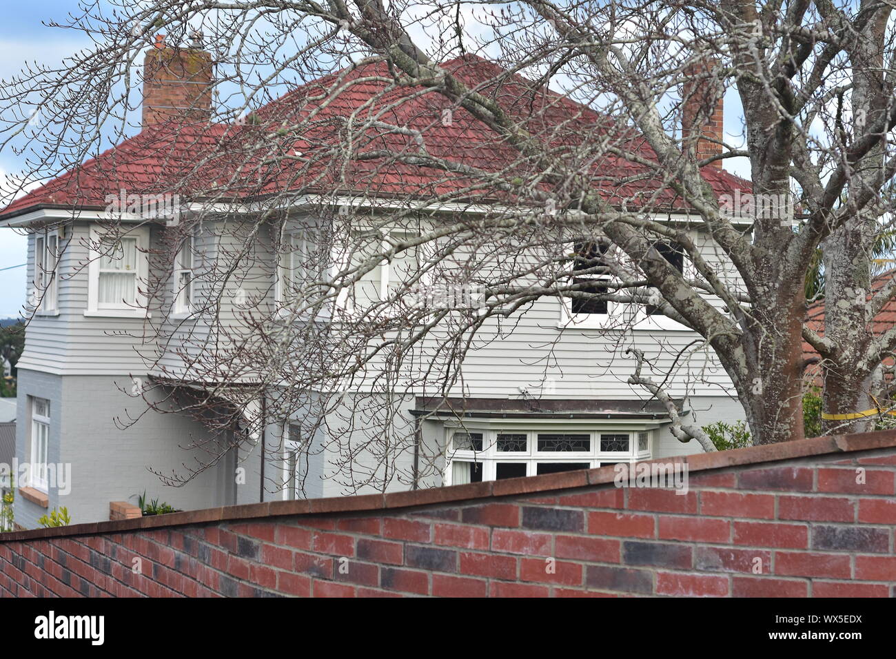 Große hölzerne Haus mit zwei Schornsteinen hinter blattlosen Baum und roten Backstein Zaun. Stockfoto