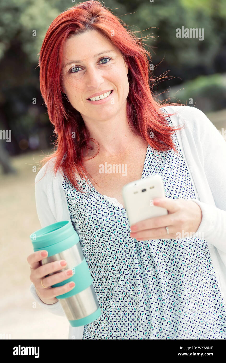 Schönen Lächeln rothaarige Frau mit einem Handy mit einer Hand und Thermoskanne in der anderen Hand mit dem Hintergrund grün Stockfoto