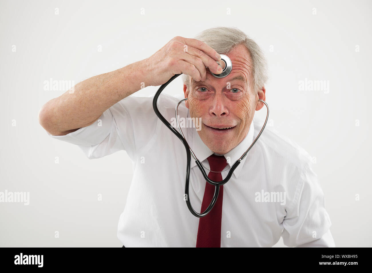Humorvoll medizinisches Konzept eines älteren Menschen ziehen ein komisches Gesicht eine stethosope auf die Stirn, als ob sein Gehirn Funktion prüfen, Intellekt, Stockfoto