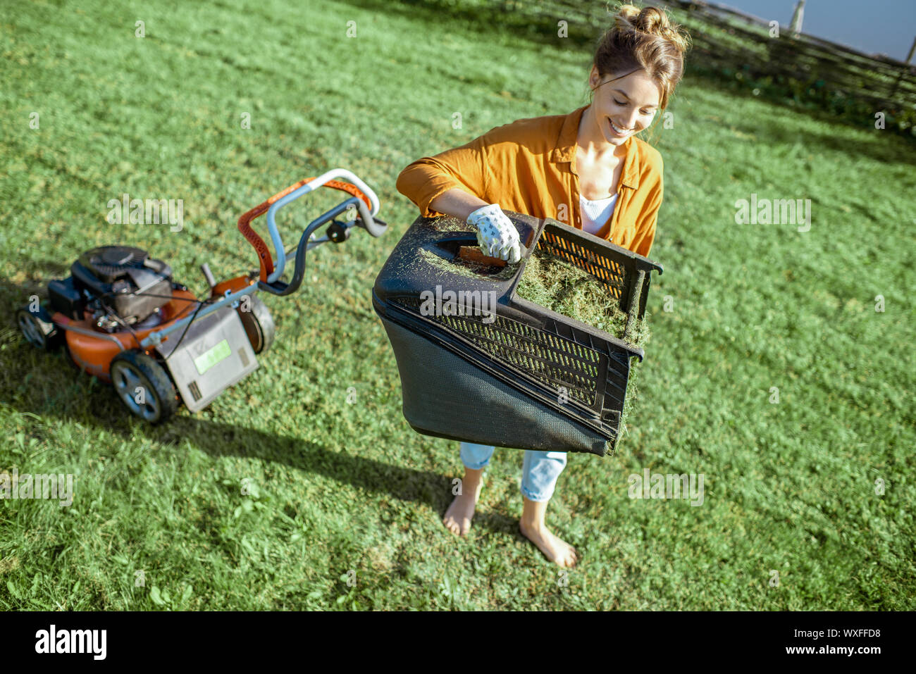 Schöne junge Frau mit Korb voller Gras bei der Gartenarbeit mit Rasenmäher auf dem Hinterhof in der Landschaft Stockfoto