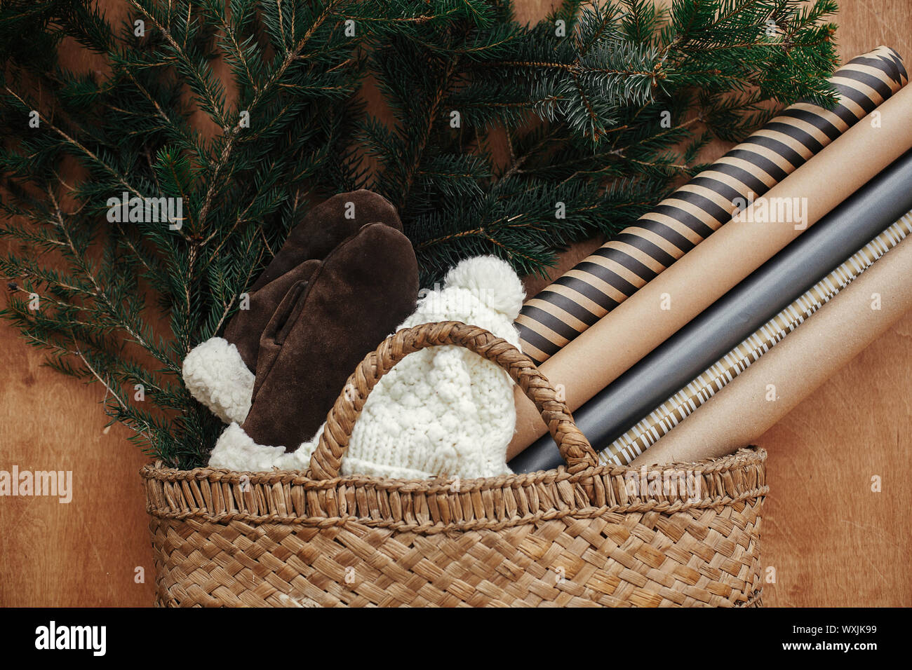 Rustikalen Korb mit Tannenzweigen, festliches Geschenkpapier, gemütliche Handschuhe und Mütze auf rustikalen Holzmöbeln Hintergrund. Flach. Winter Urlaub Vorbereitungen. Langsam l Stockfoto