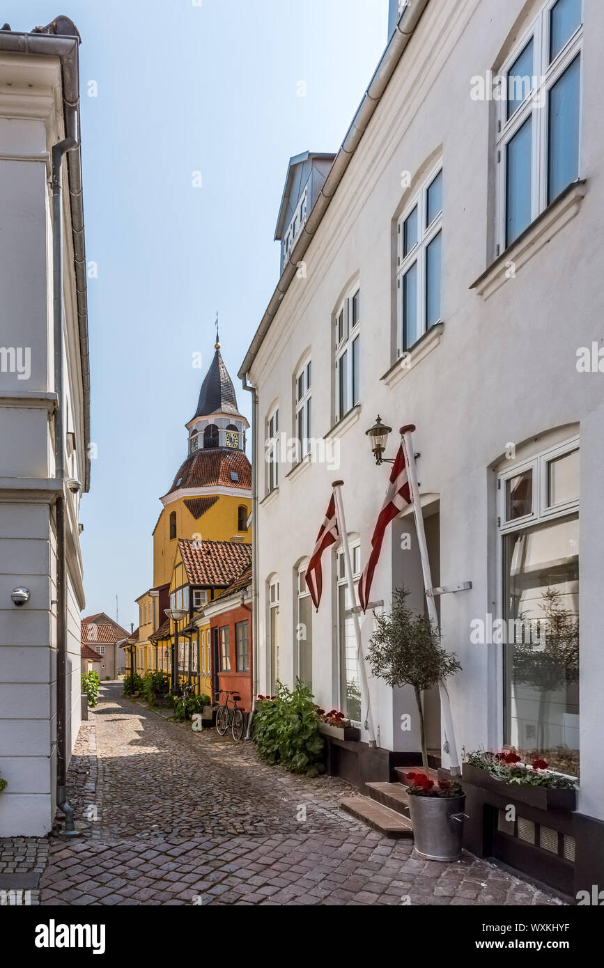 Zwei dänische Fahnen auf der Eintritt in eine schmale Gasse und der Kirchturm im Hintergrund, Faaborg, Dänemark, 12. Juli 2019 Stockfoto