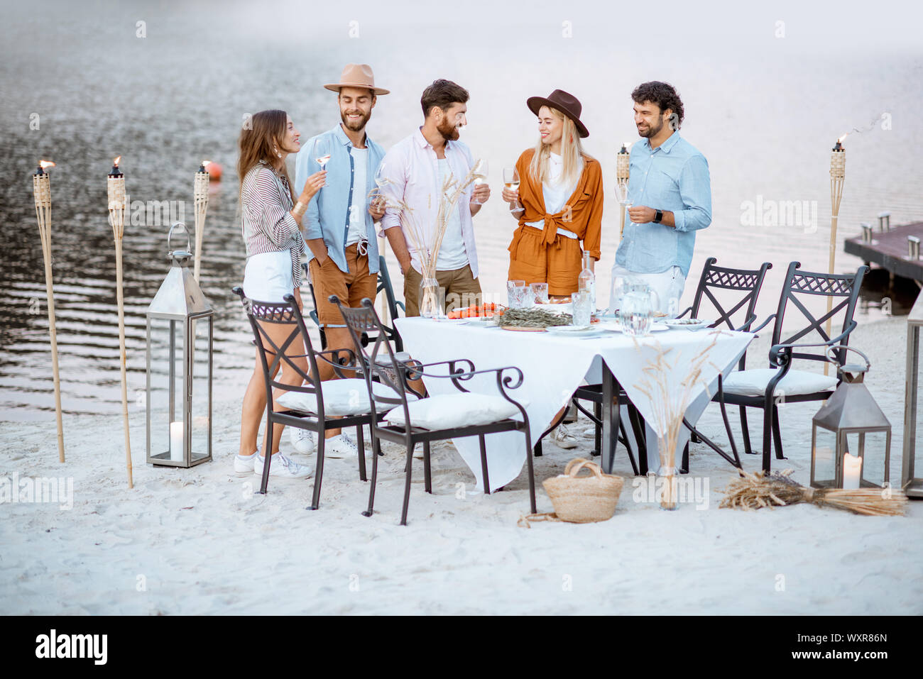 Junge Freunde heraus zusammen hängen, stehend mit Weingläsern in der Nähe der wunderschön dekorierten Esstisch am Strand bei Sonnenuntergang Stockfoto