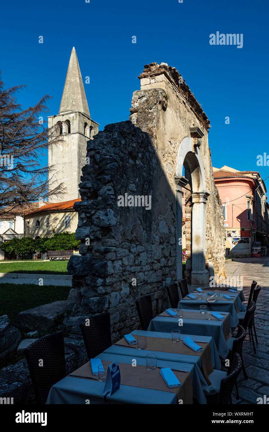 Blick auf die Glockentürme, Bruchstücke der Mauer mit einem bogenförmigen Durchgang in der Mauer und Restauranttische. Porec Parenzo, Istrien, Kroatien 2019 Stockfoto