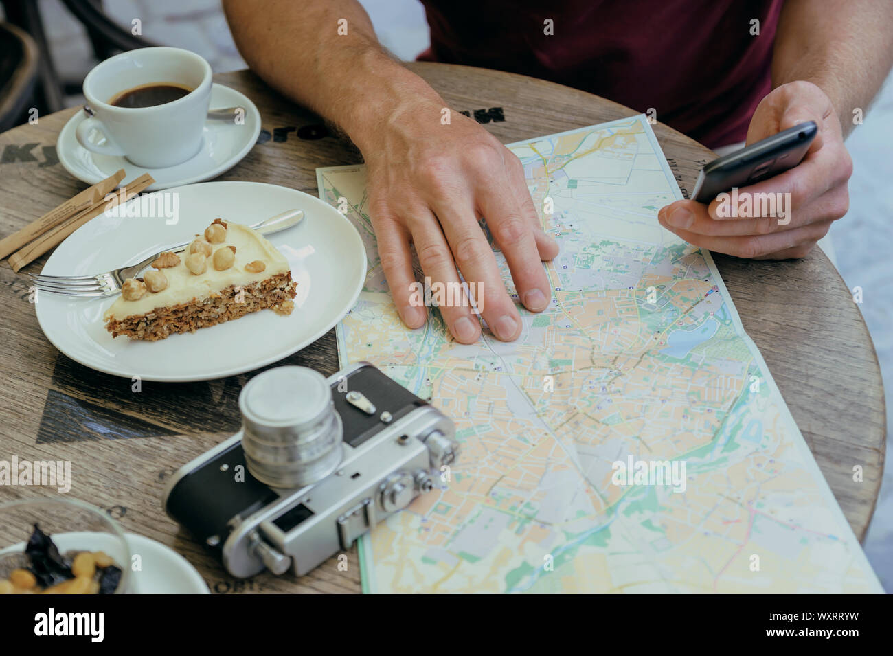 Ein Mann hält ein Smartphone. Er sitzt im Cafe Tabelle auf der Straße. Auf dem Tisch gibt es eine alte Kamera, eine touristische Karte, einen Kaffee und ein Pi Stockfoto