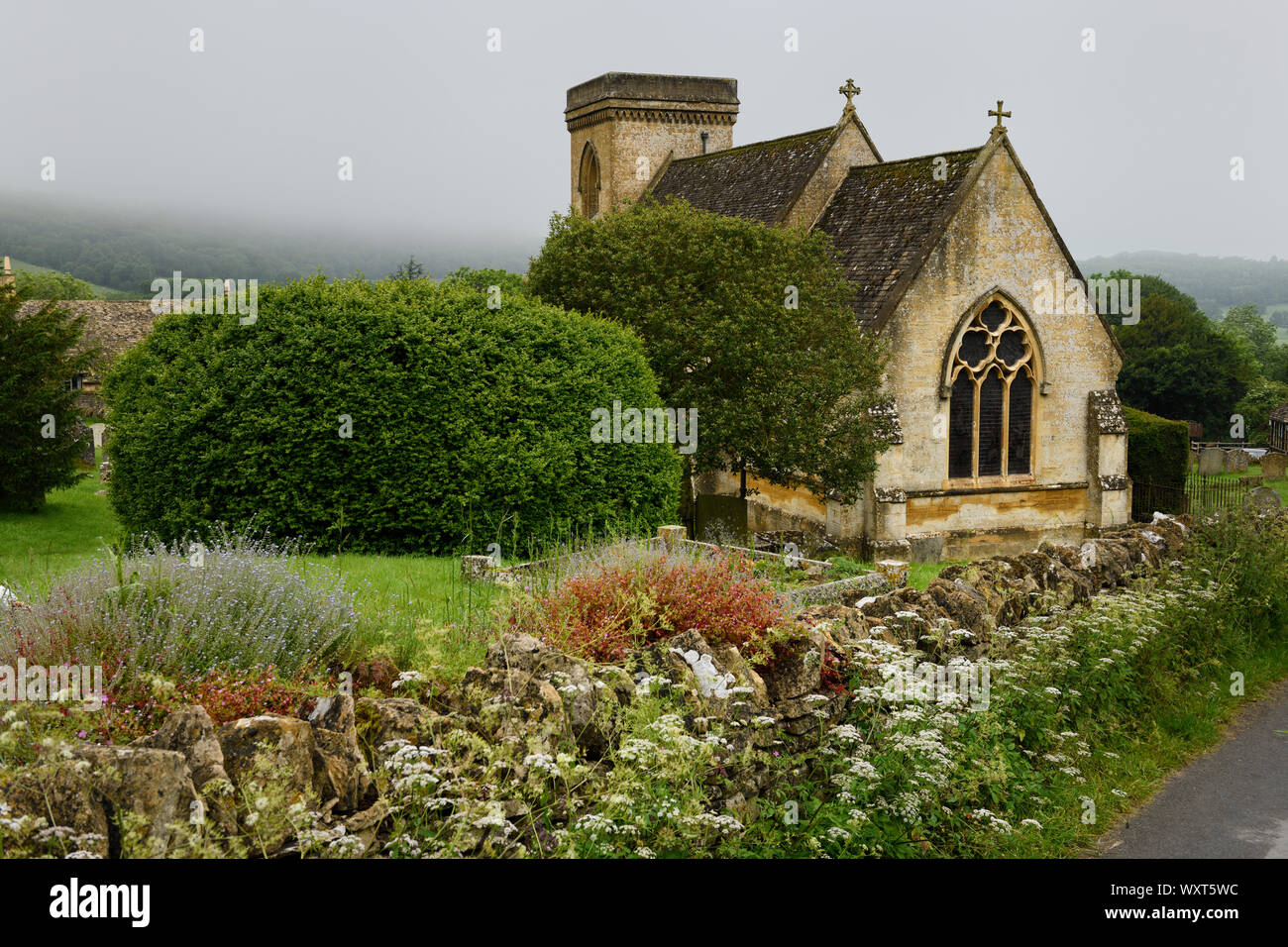 St Barnabas anglikanische Kirche mit Friedhof und Wildblumen an der Steinmauer in feuchten regnerischen Wetter in Snowshill Cotswold England Stockfoto