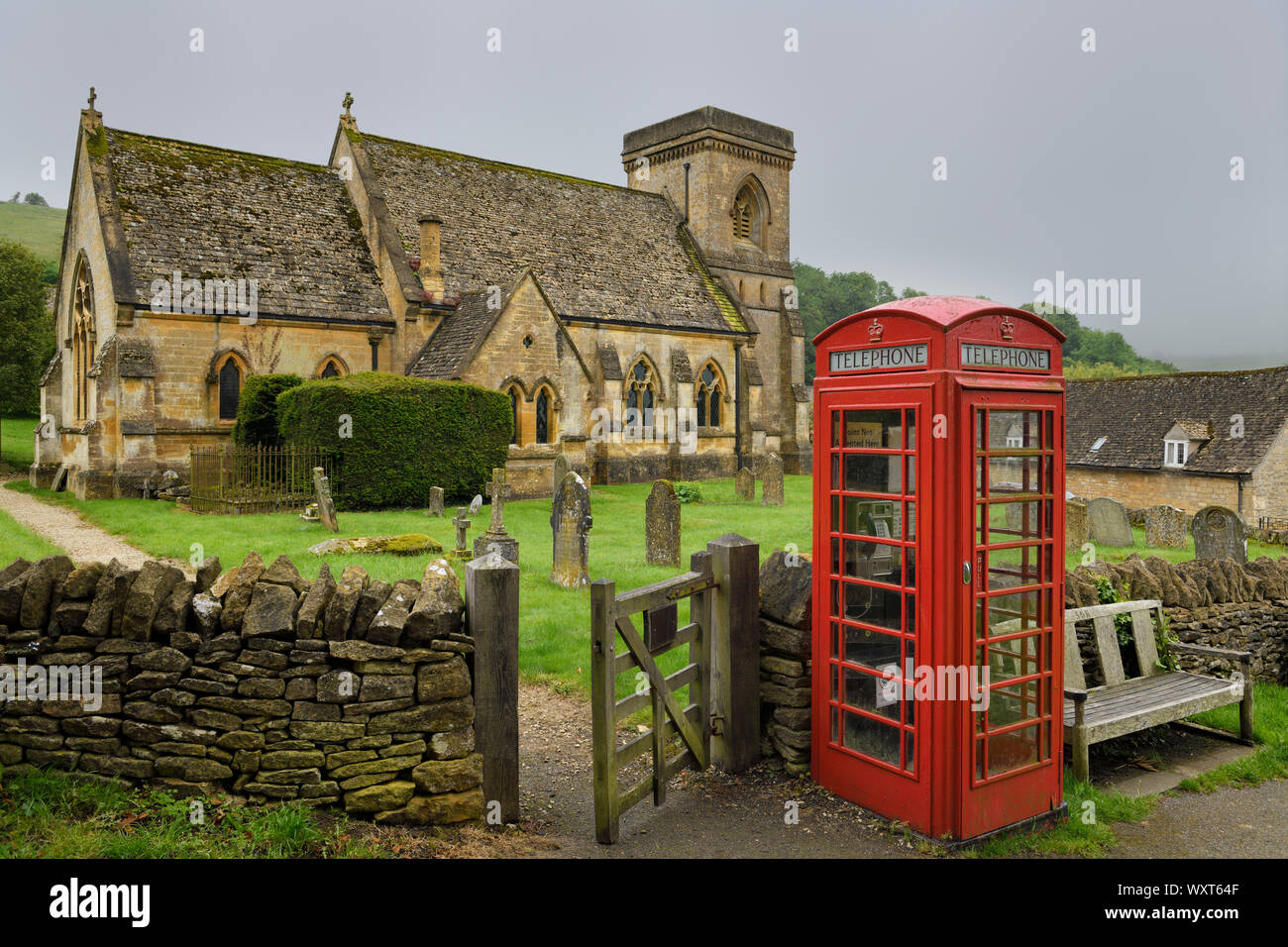 Rote Telefonzelle im 15. Jahrhundert der hl. Barnabas anglikanische Kirche mit Friedhof und Mauer Tor in feuchten regnerischen Wetter in Snowshill Cotswold England Stockfoto