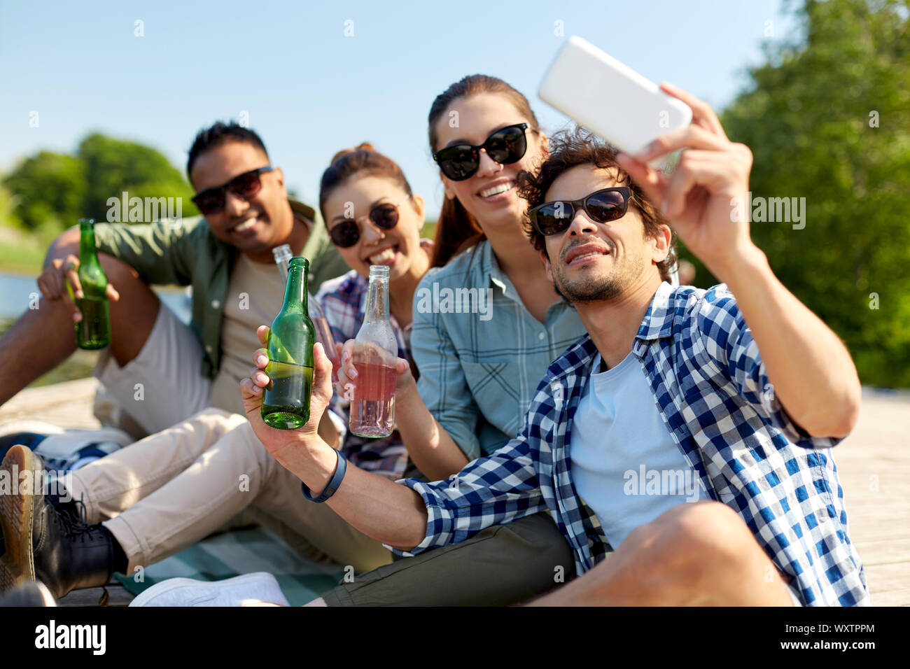 Freizeit-, Picknick- und Technologiekonzept - Freunde mit Getränken unter Berücksichtigung von selfie Smartphone auf See Pier im Sommer Park Stockfoto