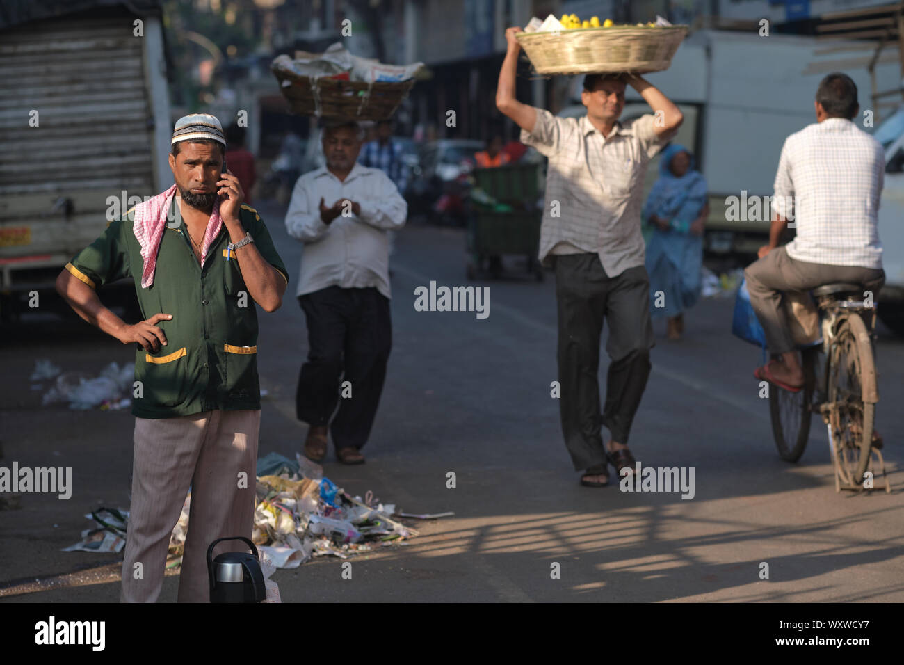 Ein moslemischer Mann in einem Markt, in Mumbai, Indien, mit seinem Handy, während hinduistische Wanderarbeiter Torhüter aus Nordindien vorbei Stockfoto