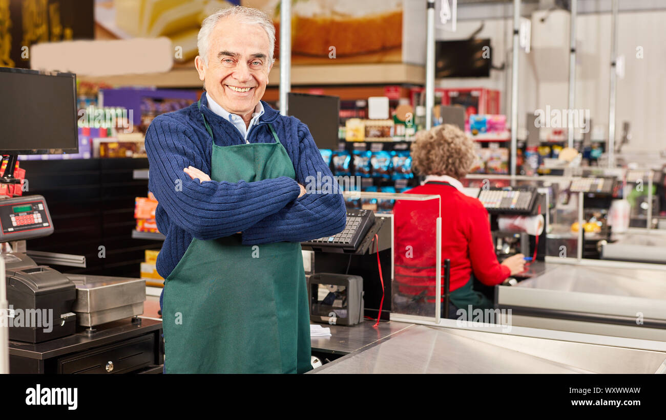 Senior als Kassiererin steht mit verschränkten Armen an der Supermarkt Kasse Stockfoto