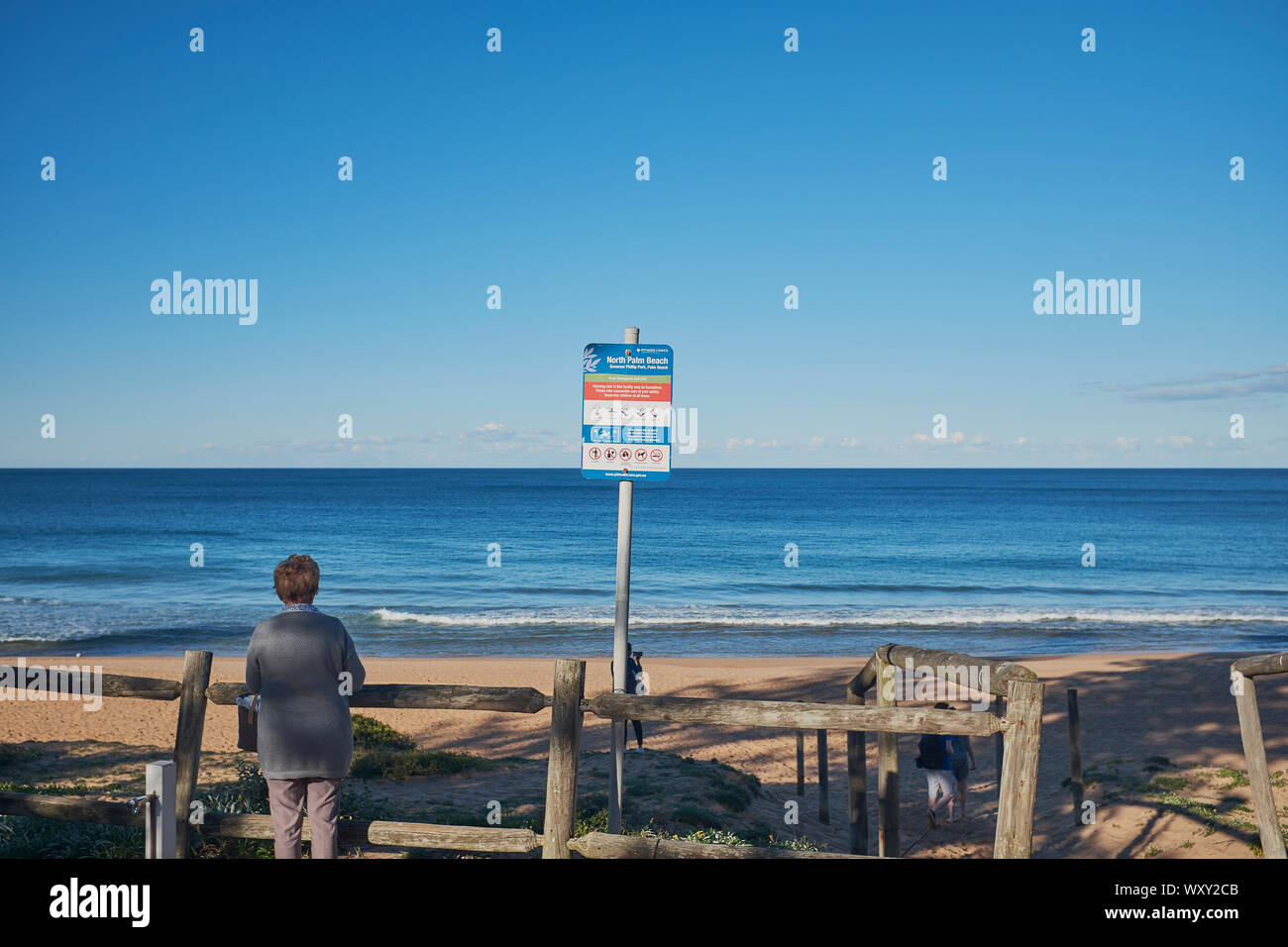 Eine Frau neben einem hölzernen Zaun am Eingang zum Palm Beach mit einem Warnschild an einem sonnigen Tag mit einem klaren blauen Himmel, New South Wales, Australien Stockfoto