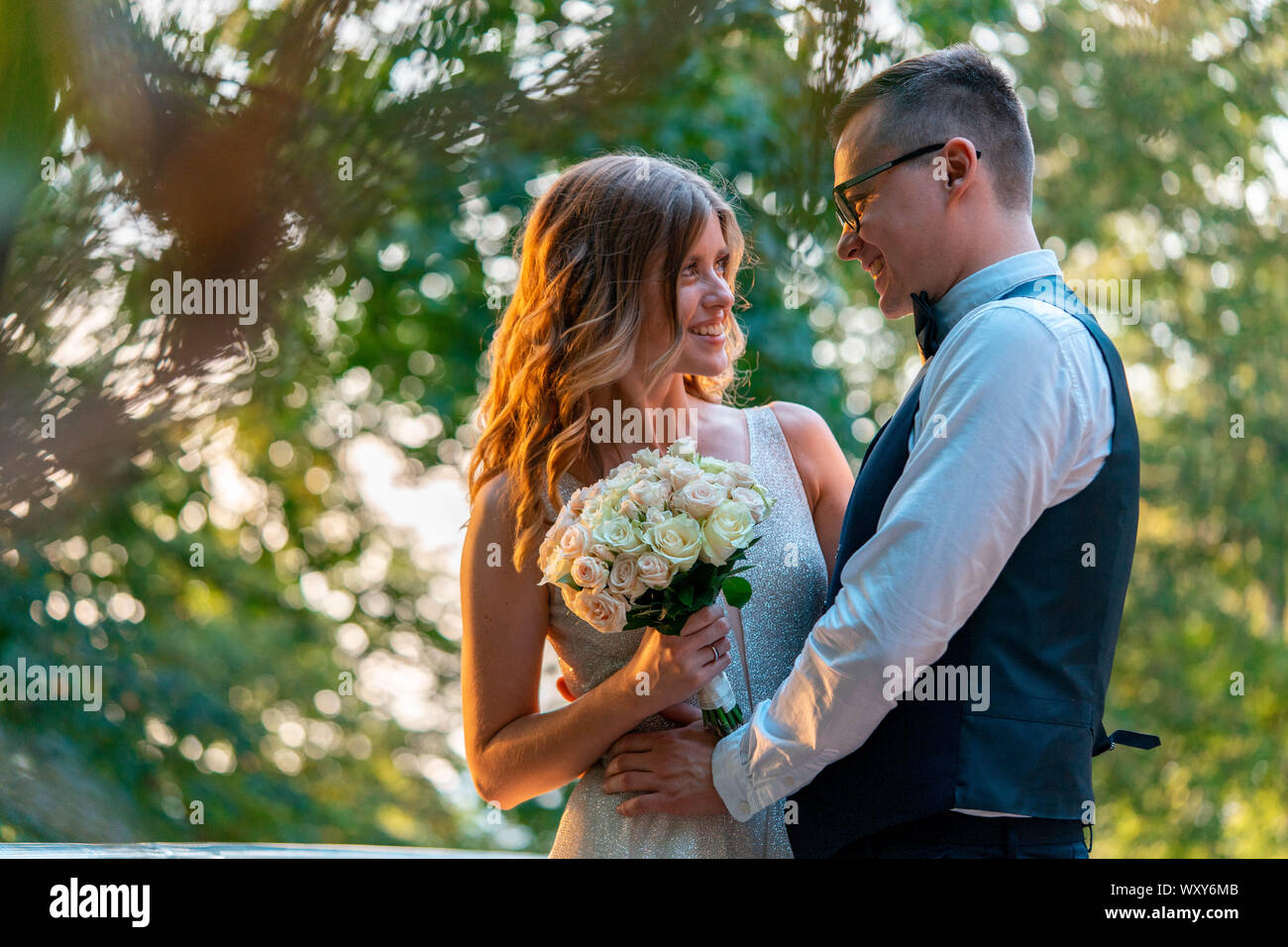 Frisch vermählte Paar stehen eng zusammen und umarmen, mit einem Lächeln. Sommer Park mit schönen Sonnenuntergang ist auf Hintergrund und verschwommen. Stockfoto