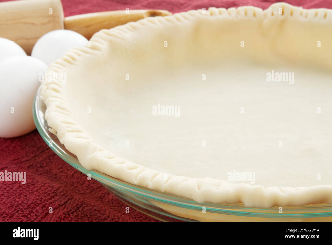 Leere ungekochten Teig Torte Kruste mit bogenkante in einem Glas Torte Platte. Es gibt weissen rohe Eier und eine hölzerne Rolling Pin im Hintergrund. Stockfoto