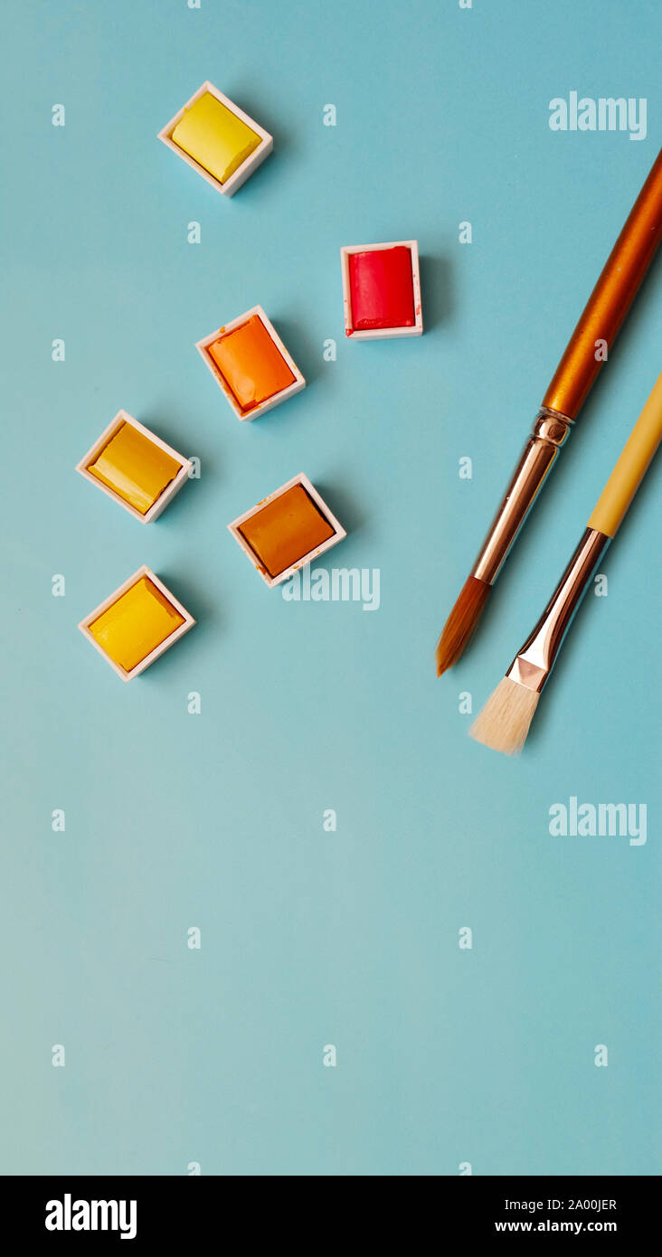 Pincel y paletas de colores cálidos de rojo, naranja y amarillo, con fondo azul. Foto de stock