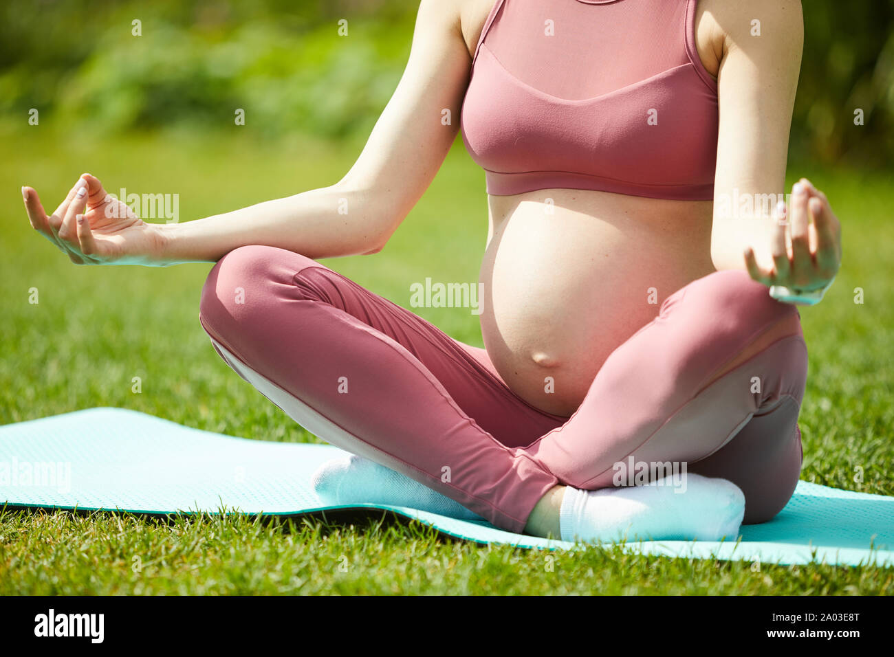 Close-up de joven embarazada en ropa deportiva rosa sentado en colchoneta de ejercicios en la postura del loto y meditando Foto de stock