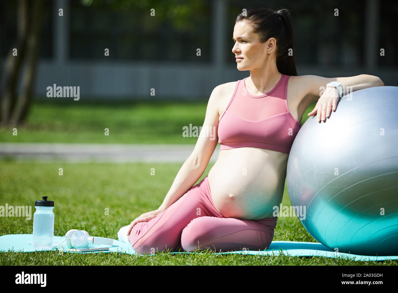 Joven embarazada con el cabello oscuro y ropa deportiva en rosa sentado en colchoneta de ejercicios con Pelota Fitness y descansando al aire libre Foto de stock