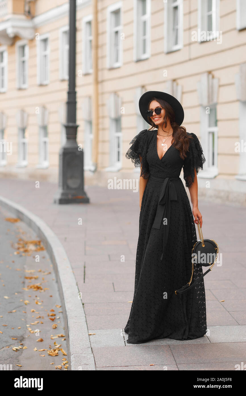 Mujer elegante en Vestido largo negro, sombrero y lentes de sol sujetando  el bolso de pie y posando en las calles de la ciudad. Elegante moda otoño  cálido mirar. L completo Fotografía